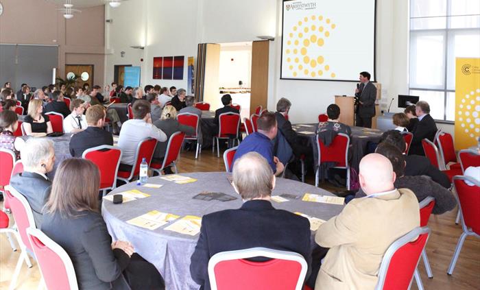 Medrus Mawr Conference Room, delegates sat at round tables viewing a presentation screen.